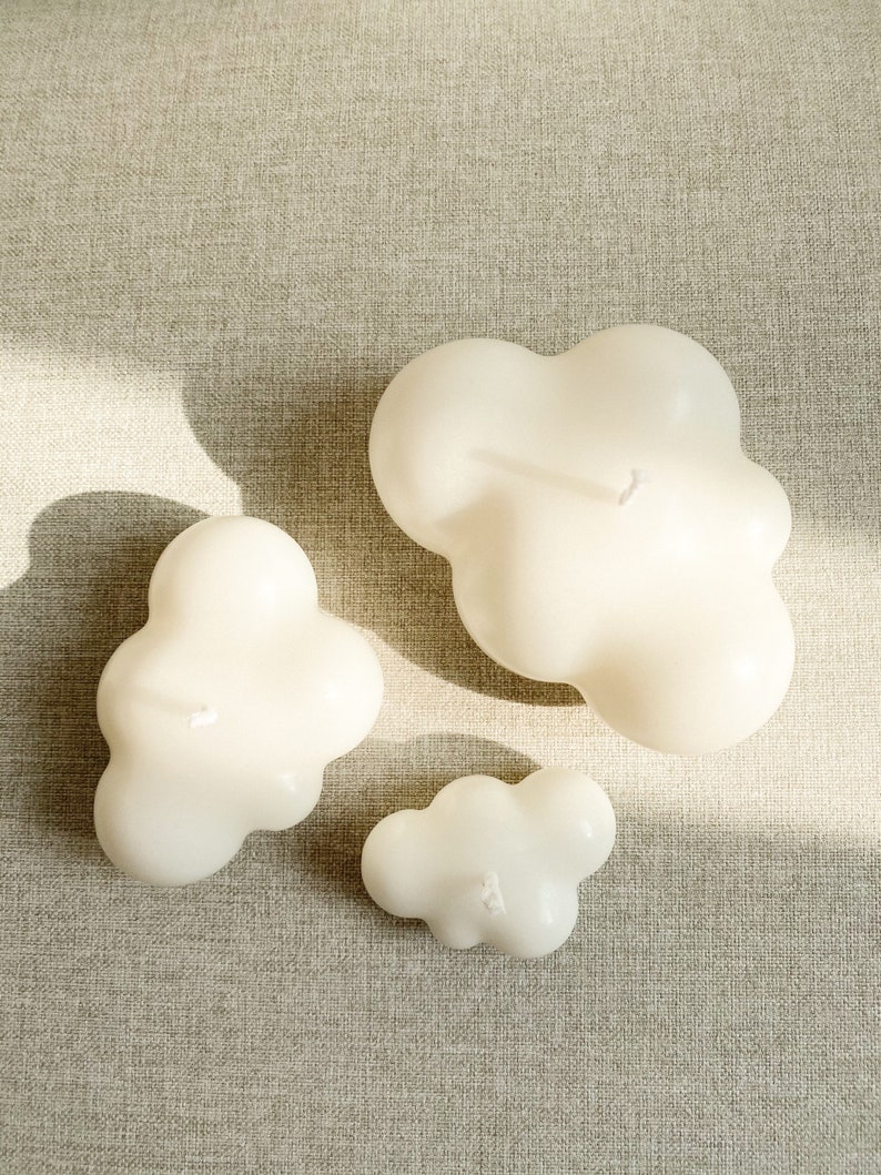May include: Three white cloud-shaped candles of different sizes on a beige textured surface.