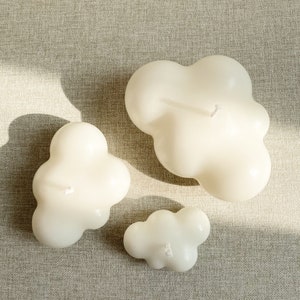 May include: Three white cloud-shaped candles of different sizes on a beige textured surface.