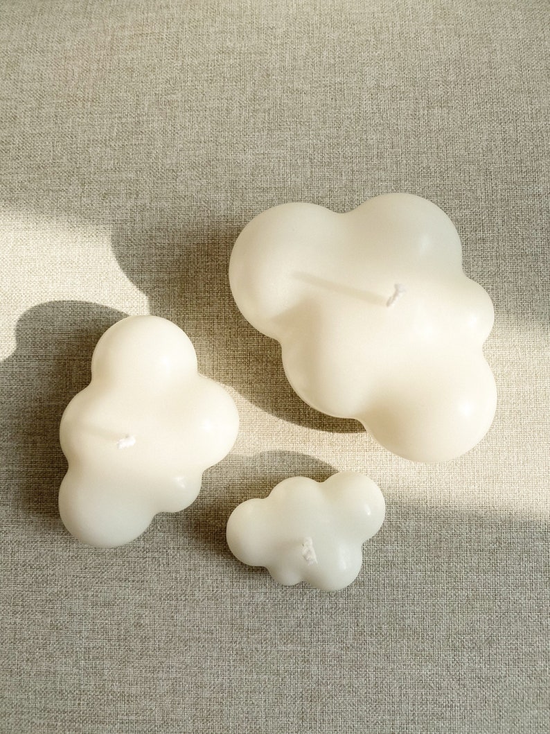 May include: Three white cloud-shaped candles of different sizes on a beige textured surface.