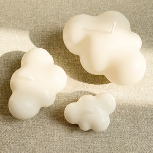 May include: Three white cloud-shaped candles of varying sizes on a beige textured surface.