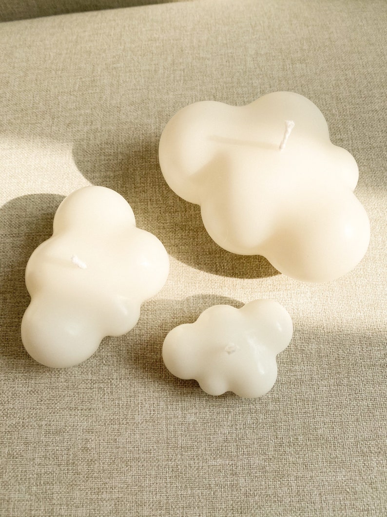 May include: Three white cloud-shaped candles of varying sizes on a beige textured surface.