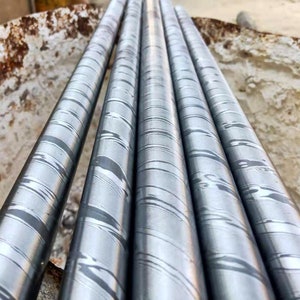 May include: Close-up of several metal rods with a unique pattern. The rods are cylindrical and have a swirling, layered design in shades of gray and silver. The rods are arranged closely together, suggesting they are part of a larger project or collection.