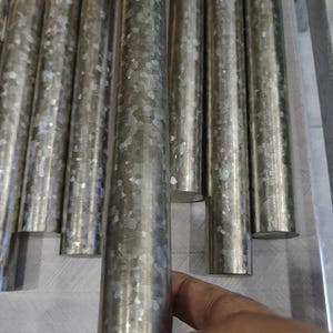 May include: Close-up of several cylindrical metal rods with a galvanized finish. The rods are arranged vertically, with one rod in the foreground being held by a hand. The metal has a mottled, speckled appearance.