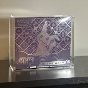 Puede incluir: Una caja de plástico transparente que contiene una caja de entrenador de élite del Juego de Cartas Coleccionables Pokémon con un diseño plateado y morado con una ilustración de Gengar. La caja tiene el texto "Escarlata & Violeta Destinos Paldea" y "Caja de Entrenador de Élite".