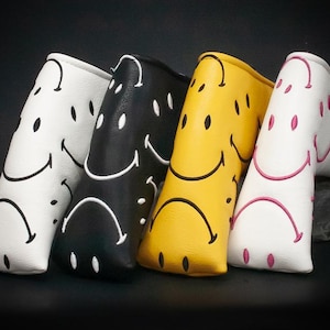 Könnte beinhalten: Vier Golfschlägerhauben in Weiß, Schwarz, Gelb und Weiß mit einem Smiley-Gesicht-Design. Die Hauben sind zum Betrachter hin abgewinkelt, mit der weißen Haube links und der weißen Haube mit rosa Akzenten rechts. Das Wort "SMILEY" ist auf der rechten Haube aufgedruckt.