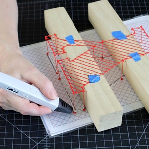 May include: A person using a 3D printing pen to create a red wireframe design on a piece of paper. The design is being held in place by two wooden blocks.