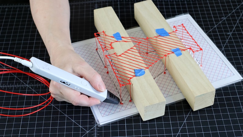 May include: A person using a 3D printing pen to create a red wireframe design on a piece of paper. The design is being held in place by two wooden blocks.