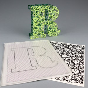 May include: A green and yellow 3D printed letter R with a circular pattern. The letter is sitting on top of a clear sheet with a grid pattern and a black and white sheet with a circular pattern.