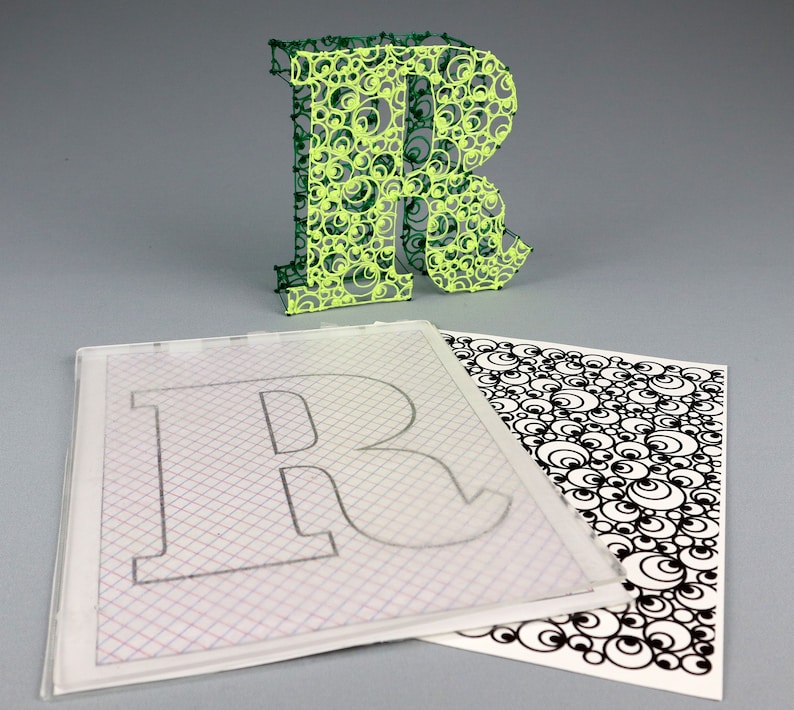 May include: A green and yellow 3D printed letter R with a circular pattern. The letter is sitting on top of a clear sheet with a grid pattern and a black and white sheet with a circular pattern.