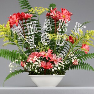 May include: A white ceramic vase with a red and green floral arrangement. The flowers are arranged in a circular pattern and the vase is sitting on a gray surface. The flowers have a "Thank You" message in white.