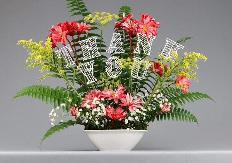 May include: A white ceramic vase with a red and green floral arrangement. The flowers are arranged in a circular pattern and the vase is sitting on a gray surface. The flowers have a "Thank You" message in white.