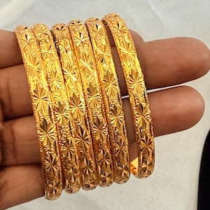 May include: Six gold bangles with a repeating starburst pattern. The bangles are a bright, shiny gold color and appear to be made of metal. The bangles are stacked together, and the image is taken from a close-up perspective.
