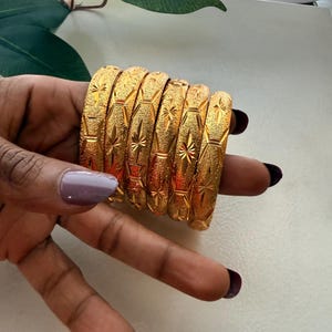May include: A set of six gold-toned bangles featuring a textured surface with repeating star-shaped engravings. Each bangle has a consistent cylindrical shape and a metallic finish that reflects light across the patterned surface of the jewelry.