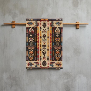 May include: A rectangular, patterned textile with a Native American design, displayed on a wooden towel rack. The textile features a cream-colored central panel with symmetrical patterns in black, red, yellow, and blue. The rack is made of light brown wood.