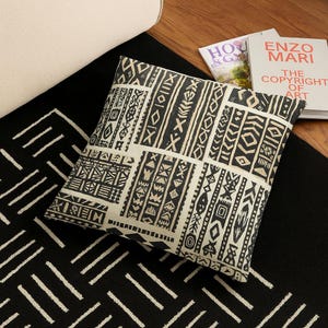 May include: A decorative pillow with a black and beige geometric pattern. The pillow is rectangular and features a variety of shapes and lines. The pillow is placed on a black and white patterned rug. Two books are visible in the background.