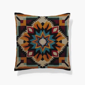 May include: Square throw pillow with a geometric pattern in shades of blue, orange, yellow, red, and black. The design features a central starburst motif surrounded by repeating shapes. The pillow has a black border.