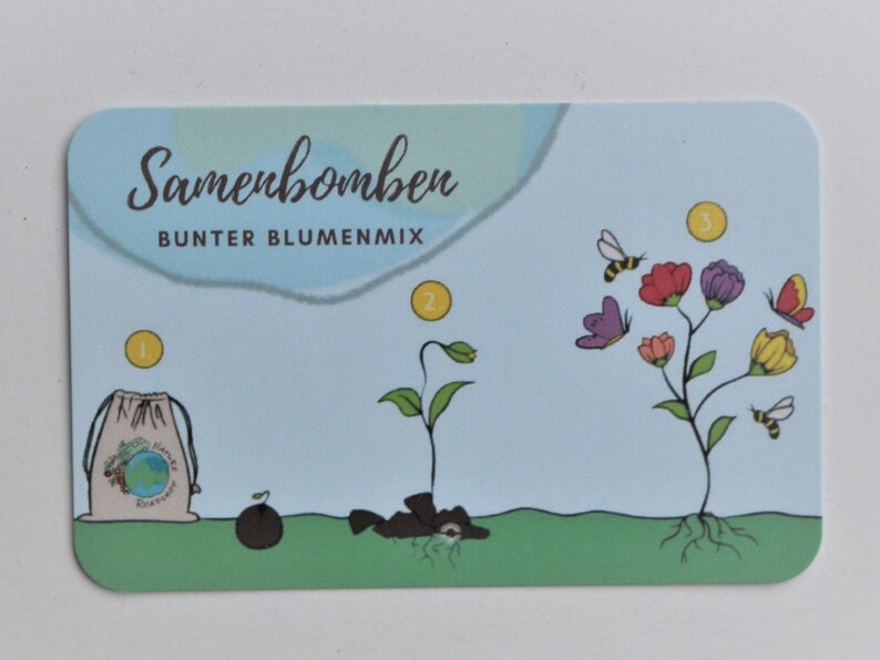 Op de afbeelding: Een rechthoekige kaart met afgeronde hoeken toont een lichtblauwe achtergrond en een afbeelding die de groei van bloemen illustreert. De kaart bevat de tekst "Samenbomber Bunter Blumenmix" en illustraties van een zaadbom, een ontkiemende plant en bloeiende bloemen met bijen en vlinders.