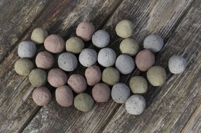 Op de afbeelding: Een verzameling kleine, ronde zaadbommen in verschillende aardetinten, waaronder grijs, roze en groen. De zaadbommen zijn gerangschikt op een verweerd houten oppervlak, wat een natuurlijke en organische uitstraling cre&euml;ert.