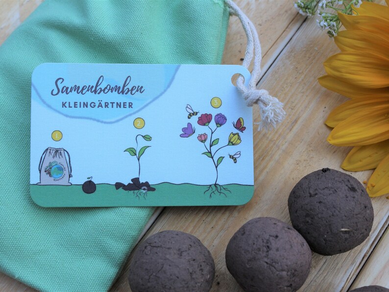 K&ouml;nnte beinhalten: Samenbomben und eine Karte mit den Worten "Samenbomben KLEING&Auml;RTNER" werden auf einer Holzoberfl&auml;che gezeigt. Die Karte zeigt eine Illustration des Pflanzenlebenszyklus. Ein gr&uuml;ner Kordelzugbeutel und eine Sonnenblume sind ebenfalls im Bild.