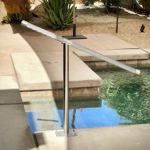 May include: A silver metal handrail with a twisted design extends over a swimming pool. The handrail is attached to a vertical post with a square base.