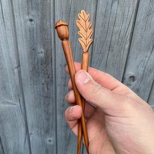 Puede incluir: Dos palos de pelo de madera con diseños tallados de bellota y hoja de roble. Los palos son marrones y tienen un acabado de madera natural.