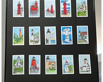May include: A framed collection of 15 watercolor paintings depicting lighthouses in various styles and settings. The paintings are arranged in a grid pattern on a black background. The frame is black and has a white mat.