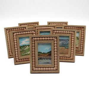 May include: A collection of small, rectangular wooden picture frames. Each frame features a landscape painting and is bordered with small, round wooden beads. The paintings depict various outdoor scenes with blue skies and natural elements.