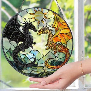 Może przedstawiać: Słoneczny łapacz wykonany ze szkła witrażowego przedstawiający dwa smoki, jeden czarny, a drugi pomarańczowy, które patrzą na siebie. Smoki otoczone są słońcem i chmurami. Łapacz słońca jest okrągły i ma metalowy łańcuch do zawieszenia.