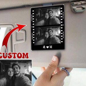 May include: A black and white photo of a couple in a car sun visor frame. The frame has a film strip design and the initials "A & H" with a heart in between.