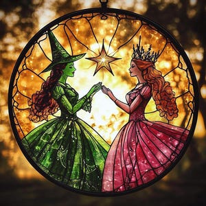 May include: Stained glass window art featuring two women, one in a green witch costume and the other in a pink princess gown, reaching for a golden star. The artwork is set in a circular black frame with a warm, golden background.