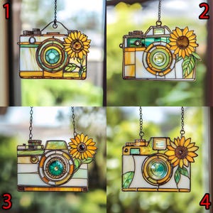 May include: Stained glass camera suncatcher with a sunflower accent. The camera features a yellow, green, and white colour scheme, with a chain for hanging. The sunflower is yellow and brown with green leaves. A decorative item for home decor.