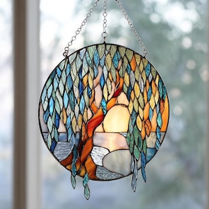 May include: Round stained glass window hanging with a tree design. The tree has a red trunk and branches, with blue, green, and yellow leaves. The background is a mix of colors, including white, orange, and blue. The window is suspended by a metal chain.