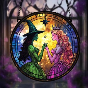 Peut inclure: Une fenêtre en verre coloré suspendue représente deux femmes, l'une en robe verte et chapeau de sorcière, l'autre en robe rose et couronne. Elles se tiennent la main devant un cadran d'horloge avec des étoiles et un fond de galaxie.