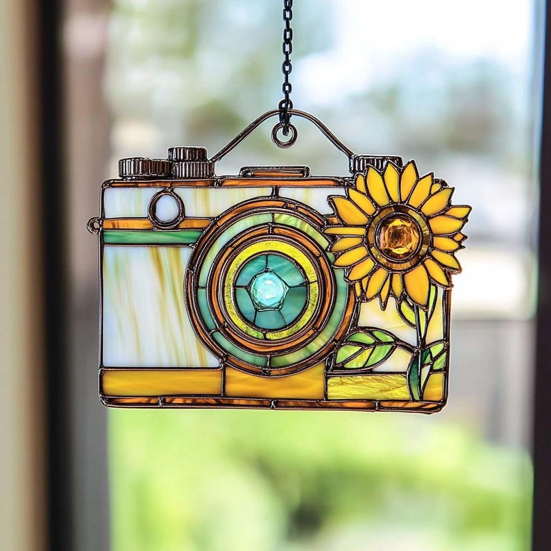 May include: Stained glass camera with a sunflower design. The camera has a yellow, green, and white colour scheme with a detailed lens and a decorative sunflower on the side. It hangs from a metal chain.