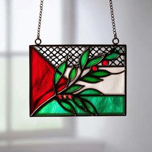 Könnte beinhalten: Rechteckige Glasmalerei mit rotem Dreieck, weißem Streifen und grünem Streifen. Ein Zweig mit grünen Blättern und roten Beeren ist abgebildet. Die Tafel ist schwarz gerahmt und hängt an einer Kette.