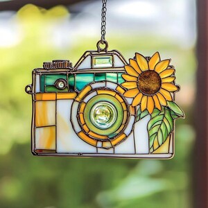 May include: Stained glass camera-shaped suncatcher with a sunflower design. The camera features yellow, green, and white glass panels, with a sunflower on the side. It hangs from a chain, ideal for window or garden decor.