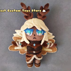 May include: A plush toy of a character with brown antlers, light brown hair, blue eyes, and a brown and beige outfit. The character is wearing a brown and blue patterned dress and has wings made of brown and beige fabric.