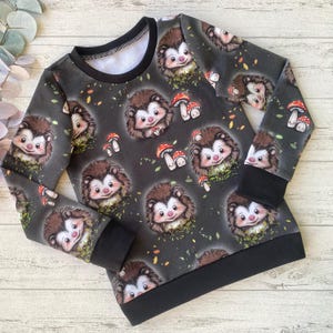 Könnte beinhalten: Schwarzes Langarmshirt mit einem sich wiederholenden Muster aus Igeln und Pilzen. Die Igel sind braun mit weißen Gesichtern und roten Nasen. Die Pilze sind rot mit weißen Punkten.