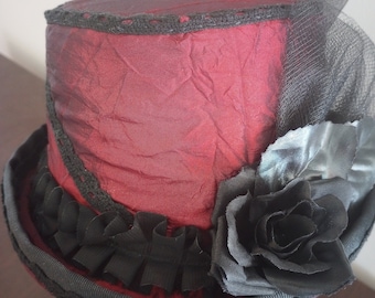 May include: A red top hat with a black lace trim and a black rose accent. The hat is made of a textured fabric and has a black tulle veil.