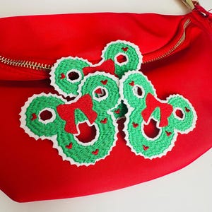 May include: Red crossbody bag with a zipper closure and two green and white embroidered Christmas wreath Mickey Mouse head patches.
