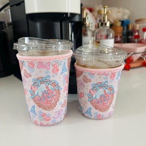 May include: Two clear plastic cups with iced coffee, each in a pink fabric sleeve. The sleeves feature a repeating pattern of castles, bows, and flowers. A white and black coffee machine is in the background.