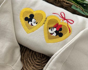 May include: A beige sweatshirt with a yellow heart-shaped locket embroidered with Mickey and Minnie Mouse. The locket is open and the two characters are looking at each other.