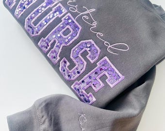May include: A gray sweatshirt with the words "REGISTERED NURSE" in large, sequined purple letters. The name "Kaitlyn" is embroidered in cursive on the sleeve. The sweatshirt is folded, with the sleeve and text visible.