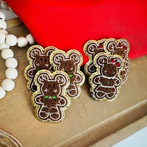May include: A set of six gingerbread man-shaped iron-on patches with gold glitter trim. The patches are brown with white icing details and green and red candy accents.