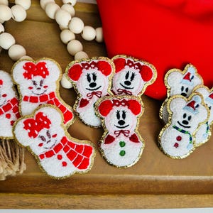 Puede incluir: Parches bordados con personajes de dibujos animados con atuendos festivos. Los parches son blancos con detalles rojos y dorados. También se ve una riñonera roja y una cadena de cuentas blancas.