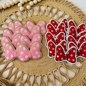 May include: A set of pink and red fabric bow appliques with white polka dots. The bows are arranged on a woven straw mat.