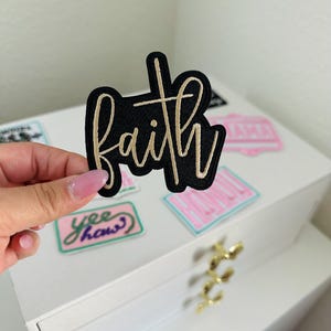 May include: Black fabric patch with the word "faith" written in gold embroidery.