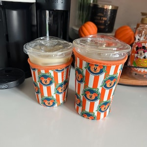 Puede incluir: Dos vasos de plástico transparente para café helado con tapa, cada uno dentro de una funda de tela con rayas verticales naranjas y blancas y motivos circulares de una calabaza con forma de Mickey Mouse y un tallo verde.