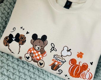 May include: A cream-colored crewneck sweatshirt featuring embroidered designs of leopard print mouse ears, a brown teddy bear wearing an orange and white checkered shirt, a coffee cup with a mouse-shaped steam, and two orange pumpkins.