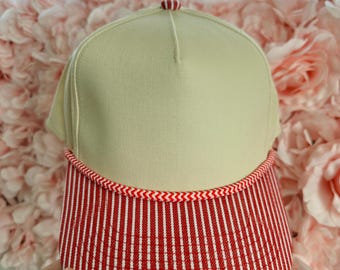 May include: A five-panel baseball cap featuring a solid beige front crown and a red and white vertical pinstripe brim. The cap includes a red and white chevron patterned rope trim detail where the crown meets the brim and a small red button.